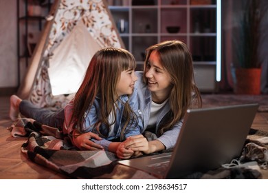 Caucasian Pretty Blond Sisters Lying On Floor Outside Wigwam While Using Laptop For Watching Movie During Evening Leisure Time. Cozy Home And Family Concept.