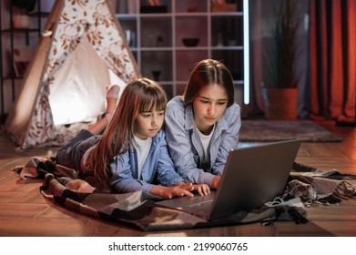 Caucasian Pretty Blond Sisters Lying On Floor Outside Wigwam While Using Laptop For Watching Movie During Evening Leisure Time. Cozy Home And Family Concept.