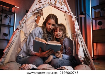Caucasian pretty blond sisters sitting in wigwam and reading book during evening time. Cozy home and family concept.