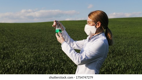 Caucasian Pretty Female Ecologist Scientist In White Gown, Mask And Googles Standing In Green Field And Looking At Chemicals In Test Tube. Woman Researcher And Biologist In Margin Studying Pesticides.