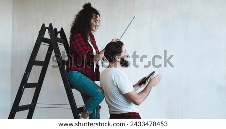 Caucasian pretty woman standing on the ladder in the room measuring wall with a measure tape and her husband coming to her with a tablet devise as showing some ideas of renovation on it.