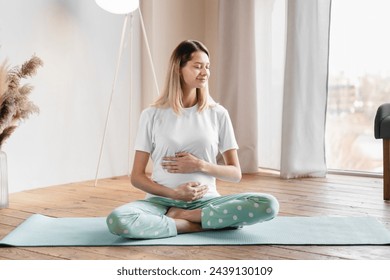 Caucasian pretty young pregnant woman holding her stomach while sitting on fitness mat in the living room. Workout, yoga and fitness at home on lockdown, pregnancy concept. Pre-natal exercises. - Powered by Shutterstock