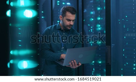 Caucasian IT professional admin using laptop computer doing data transfer operation with rack server cabinets in digital room of data center. Cyber security.
