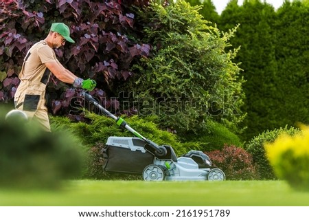Caucasian Professional Garden Worker with Cordless Electric Grass Mower Working in Residential Backyard Garden. Gardening and Landscaping Industries.