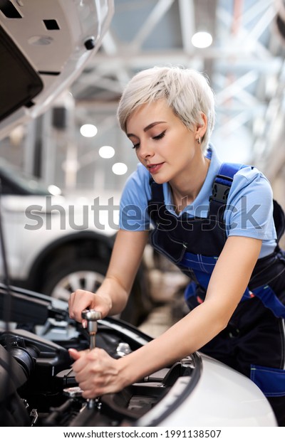 Caucasian Professional Mechanic Woman Working on\
Vehicle in Car Service. Short Haired Female Engine Specialist\
Fixing Motor, Wearing Overalls and Using a Ratchet. Modern Clean\
Workshop