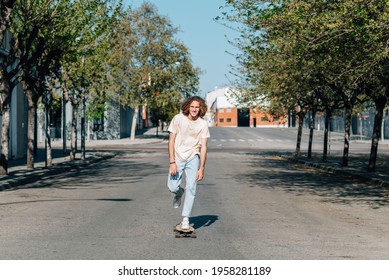 Caucasian red-haired man using a skateboard. He wears casual clothes. He skates down the road of a city surrounded by trees. Sunny summer day with harsh shadows