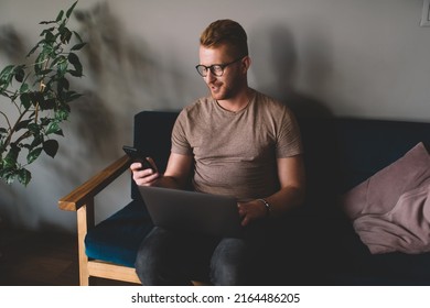 Caucasian Redhead Man Of 30 Years Old Using Smartphone App While Sitting Front Open Laptop Computer Indoors.Smiliing Young Male Reading Message On Mobile Phone During Freelance Work In Coworking Space