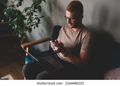 Caucasian Redhead Man Of 30 Years Old Using Smartphone App While Sitting Front Open Laptop Computer Indoors. Young Male Texting Message On Mobile Phone During Freelance Work In Coworking Space