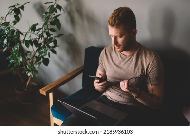 Caucasian Redhead Man Of 30 Years Old Using Smartphone App While Sitting Front Open Laptop Computer Indoors. Young Male Reading Message On Mobile Phone During Freelance Work In Coworking Spacev