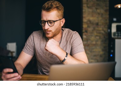 Caucasian Redhead Man Of 30 Years Old Using Smartphone App While Sitting Front Open Laptop Computer Indoors. Puzzled Male Reading Message On Mobile Phone During Freelance Work In Coworking Space