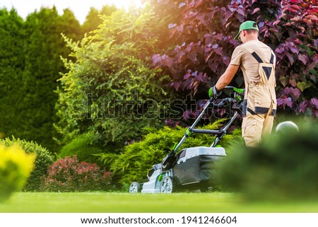 Caucasian Residential Garden Worker in His 40s Trimming Backyard Lawn Using Electric Cordless Grass Mower. Landscaping and Gardening Industry Theme.