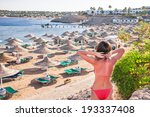 Caucasian resting girl is standing and enjoying view  of ocean and beach with parasoles. Happy relaxing beautiful young female model in red bikini. Summer vacation concept (Sharm El Sheikh, Egypt).