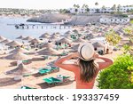 Caucasian resting girl is standing and enjoying view  of ocean and beach with parasoles. Happy relaxing female model in red bikini and white hat. Summer vacation concept (Sharm El Sheikh, Egypt).
