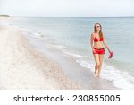 Caucasian resting girl is walking and enjoying view  of serene sea and beach. Happy relaxing female model in red bikini, shorts and with flip flop sandals. Summer vacation concept. Copy space. 