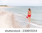 Caucasian resting girl is walking and enjoying view  of serene sea and beach. Relaxing young female model in red bikini, shorts and with flip flop sandals.  Summer vacation concept. Copy space. 