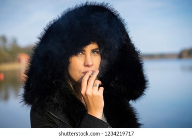 Caucasian Scandinavian Middle Age Woman Standing Outdoors In Sweden Smoking Cigarette Wearing Black Jacket And Hoodie On Head. Beautiful European Girl. Portrait Of Nice Young Woman In Nature At Lake.