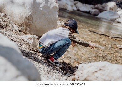 Caucasian School Boy Throwing Rocks Into The Canyon River. Kid Child Playing With Stones While Sitting On A Boulder Near Mountain River Bank.