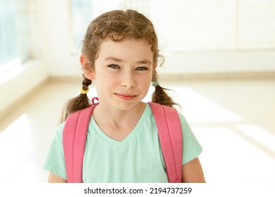 Caucasian schoolgirl with backpack stands in bright school hall with large windows. Cute caucasian girl 7 years old, elementary school student. Eye contact. Back to school, education, learning concept