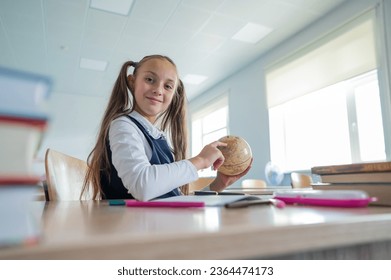 Caucasian schoolgirl sits at her desk at school and studies the globe.  - Shutterstock ID 2364474173