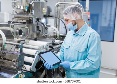 caucasian scientist man in blue lab uniform stand near manufacture machine with shafts, show empty tablet, close picture, focus on tablet, look at tablet