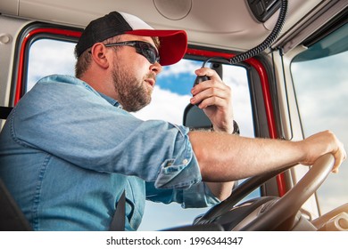 Caucasian Semi Truck Driver In His 30s Taking Conversation Using CB Radio Inside His Truck Cabin. 