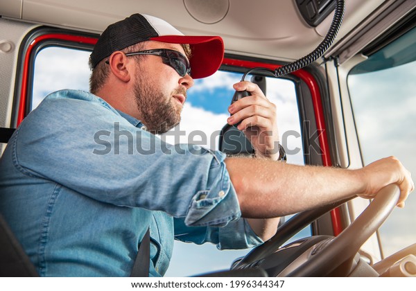 Caucasian Semi Truck Driver in\
His 30s Taking Conversation Using CB Radio Inside His Truck Cabin.\
