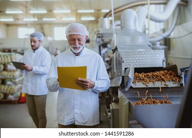 Caucasian Senior Adult Quality Controller Holding Folder With Documents And Checking On Quality Of Salty Sticks. Food Plant Interior.