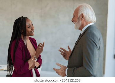 A Caucasian senior businessman exchanges ideas with a Black businesswoman, both professionally dressed, in a well-lit modern office environment. - Powered by Shutterstock