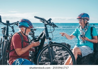 Caucasian senior cyclist couple at the beach with electric bikes in a sunny day. Old Man checks the battery while woman uses phone - Powered by Shutterstock