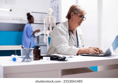 Caucasian senior doctor typing on laptop in clinical office, working on patient treatment plans. Elderly female physician with calm expression using digital technology to prepare for consultation. - Powered by Shutterstock