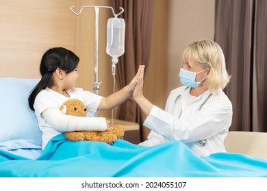 Caucasian senior doctor wear surgical mask stand check up, tease and encouragement Asian girl kid patient, Broken arm hold teddy bear sit on hospital bed to get treat and take care heal arm splint.