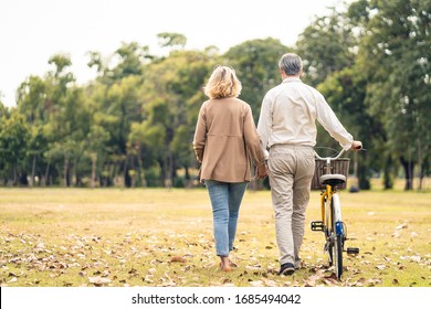 Caucasian Senior Elder Couple Love To Hold Hand And Carry Bicycle Walk In Park. Mature Are Happy And Enjoy With Slow Life. Two People Delight Activity Together. Retirement Family Lifestyle Concept.