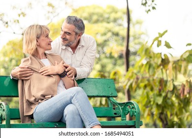 Caucasian Senior Elder Couple Sit On Bench In Park. Mature Are Happy And Enjoy With Slow Life. Old Man Love, Hug Woman From Behind. They Look And Smile Together. Retirement Family Lifestyle Concept.