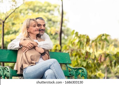 Caucasian Senior Elder Couple Sit On Bench In Park. Mature Are Happy And Enjoy With Slow Life. Old Man Love, Hug Woman From Behind. They Smile With Life Together. Retirement Family Lifestyle Concept.