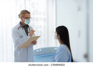 Caucasian Senior Female Doctor Explaining And Giving Advice To Asian Patient In Medical Room Together. Mature Physician And Young Woman Wear Protective Face Masks Talking Together In Hospital.