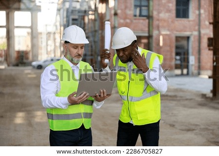 Caucasian senior foreman with wireless laptop discussing about all peculiarities of project to african american male architect at construction area. Concept of business meeting and building process