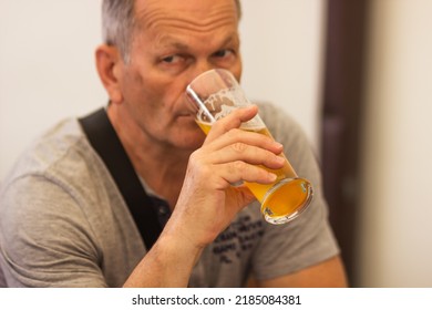 Caucasian senior man with gray hair in gray t-shirt holding and drinking glass of light beer in craft czech brewery. Alcoholic man's drink. Weekend meeting with friends in pub. Oktoberfest concept.