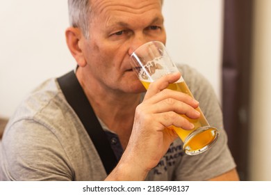 Caucasian senior man with gray hair in gray t-shirt holding and drinking glass of light beer in craft czech brewery. Alcoholic man's drink. Weekend meeting with friends in pub. Oktoberfest concept.