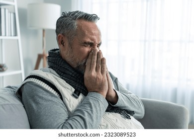 Caucasian senior man having running nose and sneezing in living room. Attractive elderly male sitting on sofa, feeling bad and suffer from allergy then put tissue cover her nose while sneez in house. - Powered by Shutterstock