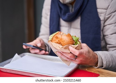 Caucasian Senior Man Holding Smartphone And Tasty Burger At The Both Hand While Eating