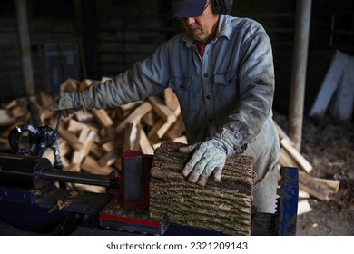 Caucasian senior man splits wood with a hydraulic log splitter. He wears a denim work shirt, work gloves and ear protection. Defocused wood pile in background, bark texture, wood splits in his hand. - Powered by Shutterstock