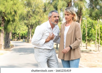 Caucasian Senior Man Suffering, Clutching And Having Chest Pain Cause From Heart Attack While Walking In The Park With His Wife, Older Woman Care And Support Husband, Health Problem And Insurance 