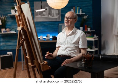 Caucasian Senior Man Working As Artist With Canvas And Easel For Drawing Masterpiece With Pencil. Older Artist Sitting In Artwork Studio Creating Fine Art As Creative Hobby Indoors