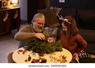 Caucasian senior man and young girl making a Christmas wreath together, smiling and enjoying festive home atmosphere with stockings and holiday decor in background - Powered by Shutterstock