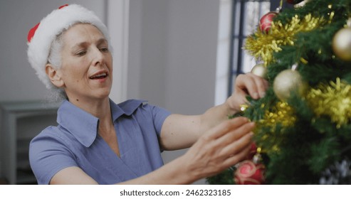 Caucasian senior woman with gray hair is decorating Christmas tree. She wears a Santa hat, has short hair, and a blue shirt - Powered by Shutterstock