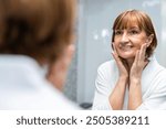 Caucasian senior woman looking at mirror then apply lotion on face. Elderly mature female in bathrobe touches on face and apply cream lotion for rejuvenation with gentle after shower. Beauty skin care