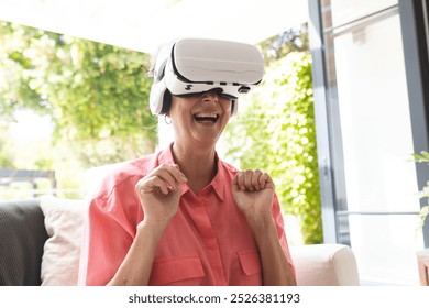 A Caucasian senior woman wearing virtual reality headset, laughing at home. Sporting short blonde hair and a pink shirt, enjoying a digital experience, unaltered - Powered by Shutterstock