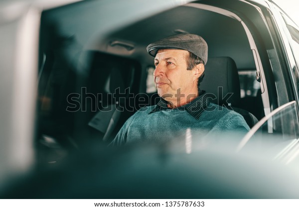 Caucasian serious senior man with cap on\
head sitting in expensive car. Window\
opened.