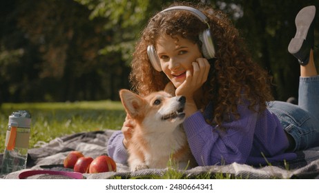 Caucasian smiling happy girl curly hair listen music in headphones relaxing summer picnic nature young pet owner handler woman rest outdoors with cute puppy welsh corgi dog best friends in park city - Powered by Shutterstock