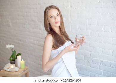 Caucasian smiling woman after spa covered with white towel holding a glass of water.